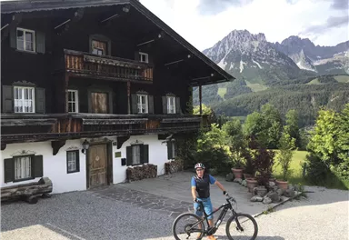 Ein Radfahrer steht vor einem traditionellen, holzverkleideten Haus in den Bergen. Im Hintergrund sind beeindruckende Berge und eine grüne Landschaft zu sehen.