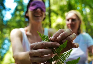 Zwei Frauen in einem Wald, eine trägt eine Augenbinde und lächelt. Die andere hält einen Farn in der Hand und schaut zu.