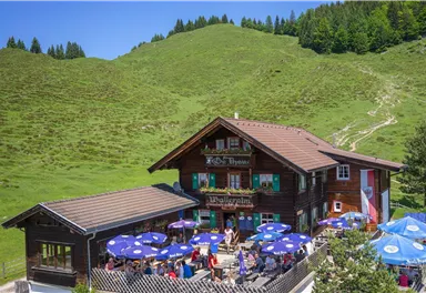 Ein malerisches Restaurant in den Bergen, umgeben von grünen Wiesen. Viele Gäste sitzen an Tischen im Freien unter blauen Sonnenschirmen.