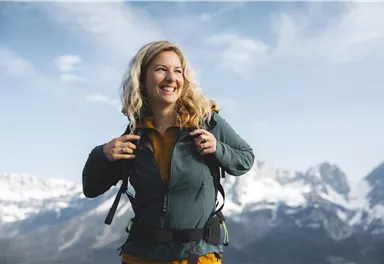 Eine lächelnde Frau mit einem Rucksack steht in den Bergen. Hinter ihr sind schneebedeckte Gipfel und ein blauer Himmel zu sehen.