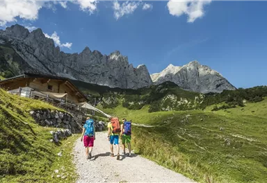 Drei Wanderer auf einem schmalen Pfad in den Bergen. Im Hintergrund sind hohe Felsen und eine malerische Landschaft zu sehen.