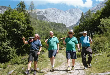 Vier Männer wandern auf einem Pfad in der Natur, umgeben von Bäumen und Bergen. Sie tragen Werkzeuge auf ihren Schultern und scheinen die frische Luft zu genießen.