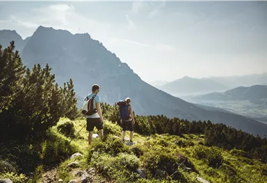Zwei Wanderer bewegen sich durch eine grüne Landschaft mit Bergen im Hintergrund. Der Himmel ist klar und die Aussicht ist atemberaubend.