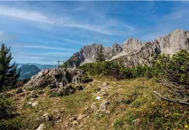Eine malerische Berglandschaft mit felsigen Gipfeln und einem kleinen Kreuz auf einem Hügel. Der Himmel ist klar und blau, umgeben von grünem Gras und Pflanzen.