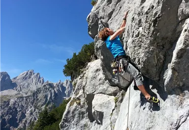 Ein Kletterer erklimmt eine steile Felswand in den Bergen. Der Himmel ist blau und die Landschaft ist von beeindruckenden Gipfeln umgeben.