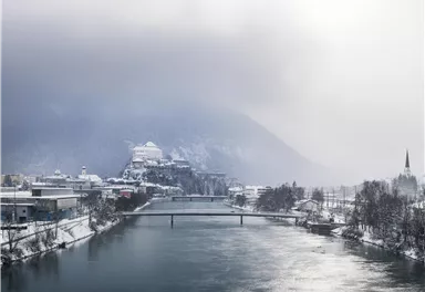 A frosty winter landscape with a peaceful river and snow-covered banks. In the background, mountains and a castle can be seen.