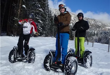 Drei Personen fahren auf Segways im Schnee. Im Hintergrund sind schneebedeckte Bäume und Berge sichtbar.