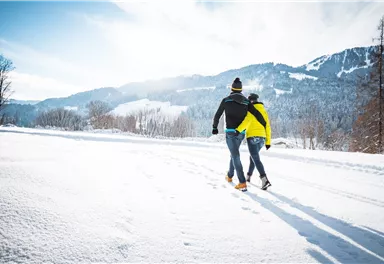 Ein Paar geht durch eine verschneite Landschaft. Die Berge sind im Hintergrund sichtbar und die Sonne scheint.