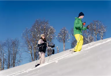 Zwei Personen wandern auf einem schneebedeckten Hang. Der Himmel ist klar und blau, umgeben von schneebedeckten Bäumen.