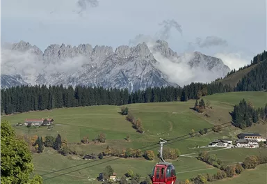 Eine Seilbahn schwebt über grüne Wiesen und kleine Dörfer. Im Hintergrund sind beeindruckende Berggipfel und Wolken zu sehen.