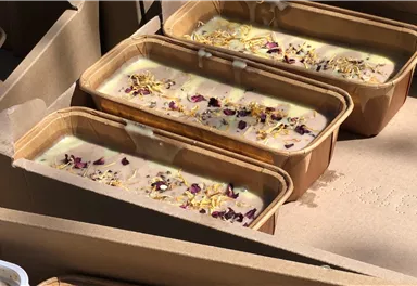 Drei Kisten mit handgemachter Seife, verziert mit trockenen Blütenblättern. Die Seife hat eine cremige Textur und schöne Farben.