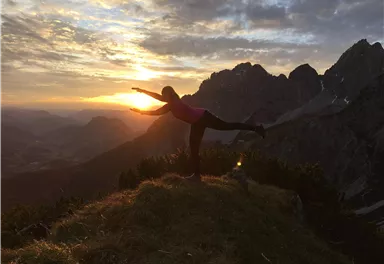 Eine Person macht eine Yoga-Pose auf einem Hügel bei Sonnenuntergang. Die Landschaft zeigt beeindruckende Berge und eine malerische Aussicht.
