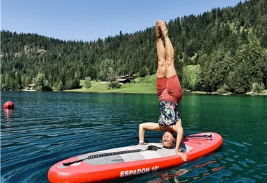 Eine Person macht einen Handstand auf einem Sup-Board in einem klaren, grünen See. Die Umgebung ist von Bäumen und Bergen umgeben, bei strahlendem Sonnenschein.