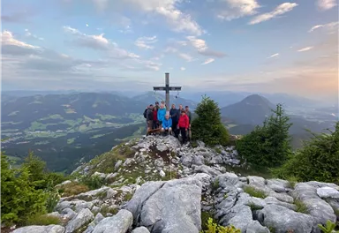 Eine Gruppe von Wanderern steht an einem Gipfelkreuz auf einem Berg. Im Hintergrund sind eine bezaubernde Landschaft und ein klarer Himmel zu sehen.