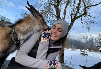 Eine Frau sitzt draußen im Schnee und lächelt, während eine Ziege sie an der Wange berührt. Im Hintergrund sind schneebedeckte Berge und Bäume zu sehen.