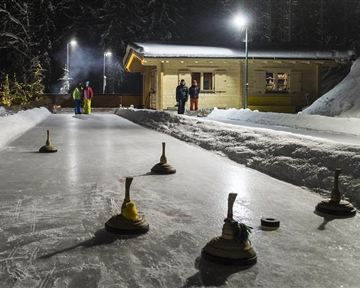 Eine Schneebahn zum Eisstockschießen bei Nacht, beleuchtet von Lichtern. Im Hintergrund stehen einige Personen und eine hölzerne Hütte.
