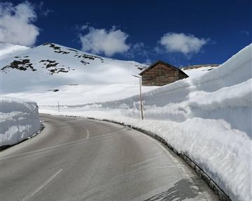 Eine schneebedeckte Straße mit hohen Schneewänden und einem Holzhaus in der Nähe. Der Himmel ist klar und blau mit einigen Wolken.
