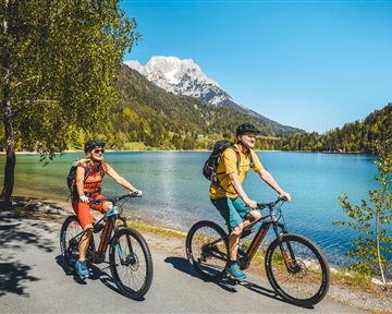Zwei Radfahrer fahren am Ufer eines blauen Sees, umgeben von grünen Bäumen und Bergen. Der Himmel ist klar und sonnig.