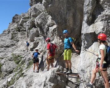 Eine Gruppe von Kletterern navigiert auf einem schmalen Pfad entlang einer felsigen Wand. Die Umgebung besteht aus beeindruckenden Felsen und einer klaren blauen Himmel.