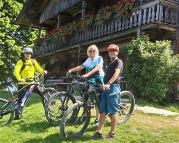 Drei Radfahrer stehen mit ihren Fahrrädern auf einer grünen Wiese vor einem holzverkleideten Haus. Die Sonne scheint und es ist ein klarer, blauer Himmel zu sehen.