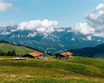 Eine malerische Berglandschaft mit grünen Wiesen und sanften Hügeln. Im Hintergrund sind majestätische Berge und Wolken zu sehen.