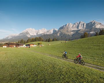 Eine schneebedeckte Straße mit hohen Schneewänden und einem Holzhaus in der Nähe. Der Himmel ist klar und blau mit einigen Wolken.