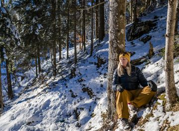A person is sitting relaxed in a snowy forest. Tall trees stand around them and the sun is shining through the branches.