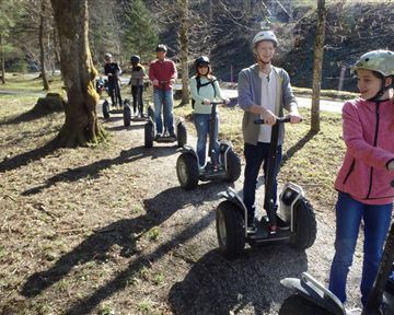 Eine Gruppe von Menschen fährt auf Segways über einen malerischen Waldweg. Die Sonne scheint und die Umgebung ist grün und einladend.