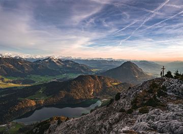 Eine beeindruckende Berglandschaft mit einem klaren Himmel und tiefgrünen Tälern. Im Hintergrund sind schneebedeckte Gipfel und ein glitzernder See zu sehen.