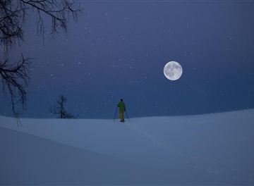 Ein Skiwanderer steht unter einem vollen Mond in einer verschneiten Landschaft. Der Himmel ist dunkelblau mit sanften Schneeflocken.