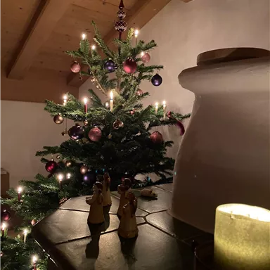 Ein festlich geschmückter Weihnachtsbaum mit Kerzen und bunten Kugeln. Im Vordergrund stehen kleine Engel-Figuren und eine brennende Kerze.