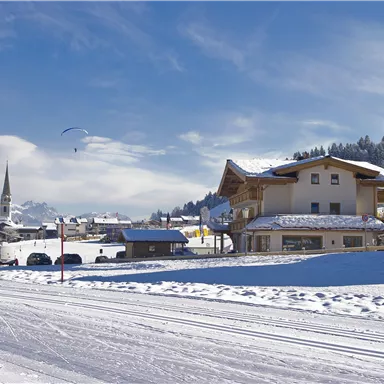 Eine malerische Winterlandschaft mit schneebedeckten Häusern und Bergen. Der Himmel ist klar mit einigen Wolken und ein Gleitschirm fliegt darüber.