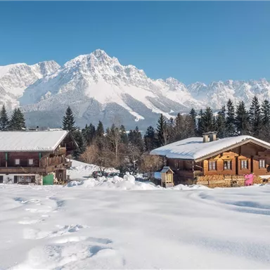 An idyllic winter scene with snow-covered cottages and majestic mountains in the background. The clear blue sky completes the peaceful atmosphere.