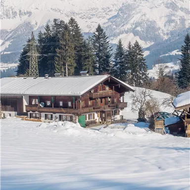 A picturesque chalet in a snow-covered landscape with mountains in the background. The surroundings are tranquil and surrounded by coniferous trees.