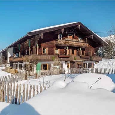 A traditional wooden house in the snow with a clear blue sky. The surroundings are covered by a white blanket of snow and a simple wooden fence.