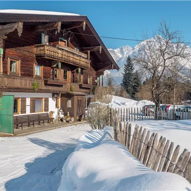 A traditional wooden house in the snow with a clear blue sky in the background. The landscape is surrounded by snow-covered trees.
