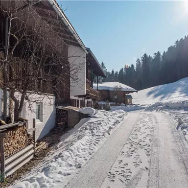 A snow-covered path leads to traditional huts in a winter landscape. In the background, snow-capped mountains and a clear blue sky can be seen.