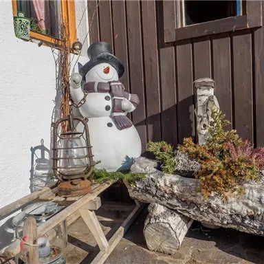 Ein dekorativer Schneemann steht neben einem Holzstamm mit festlichem Grün. Im Hintergrund sind einige alte Lampen zu sehen, die rustikale Gemütlichkeit ausstrahlen.