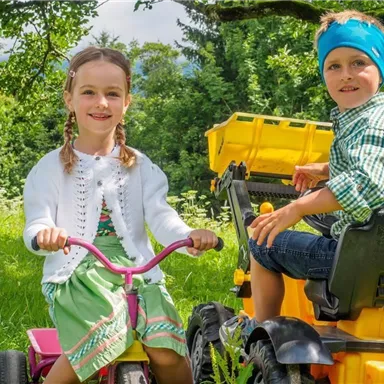 Zwei Kinder spielen im Freien, eines auf einem Bobby-Car und das andere auf einem Spielzeugtraktor. Sie lächeln fröhlich und umgeben von einer grünen Wiese mit Bäumen im Hintergrund.