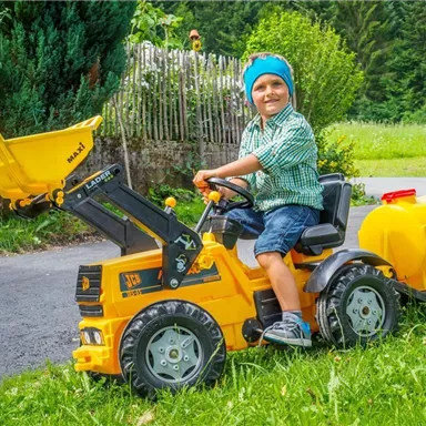 Ein fröhlicher Junge sitzt auf einem gelben Spieltraktor mit einem Anhänger. Im Hintergrund sind grüne Wiesen und Bäume zu sehen.