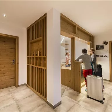 A modern reception area with a bright interior and wooden paneling. Two people are standing at a reception desk, while luggage is nearby.