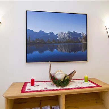 Ein gemütlicher Raum mit einem Holzregal und einer Landschaftsmalerei über einem Stillgewässer. Auf dem Regal stehen Kerzen und eine dekorative Schale.