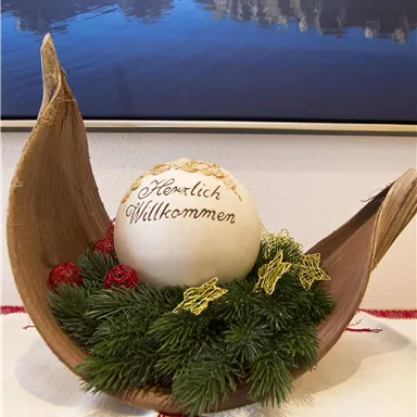 Eine dekorative Weihnachtsanordnung mit einer großen, beschrifteten Kugel in der Mitte. Umgeben von Tannenzweigen und roten Beeren, strahlt sie festliche Stimmung aus.