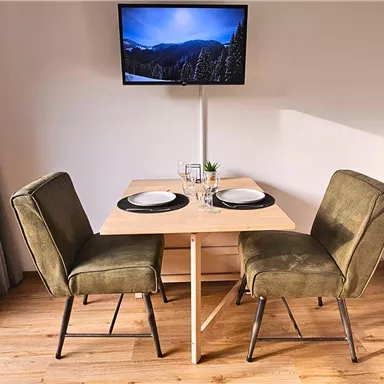 A modern dining area with a table and two green chairs. In the background, there is a television and a window through which light falls.