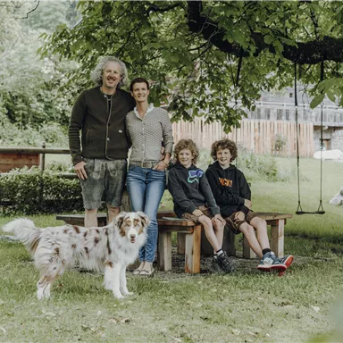Eine Familie steht im Freien unter einem Baum. Neben ihnen sitzt ein Hund und es gibt eine Schaukel im Hintergrund.