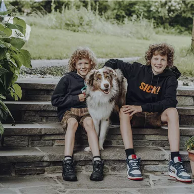 Zwei Jungen sitzen auf einer Treppe und lächeln in die Kamera. Ein Hund sitzt zwischen ihnen und es ist eine grüne Umgebung sichtbar.