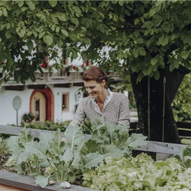 Eine Frau steht an einem Gemüsebeet und schaut lächelnd auf die Pflanzen. Im Hintergrund sind ein Haus und ein großer Baum zu sehen.
