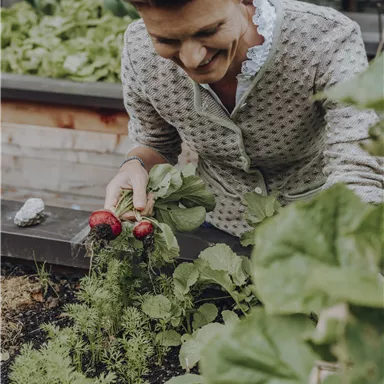 Eine Frau erntet frisches Gemüse aus einem Garten. Sie hält Radieschen in der Hand und lächelt dabei.