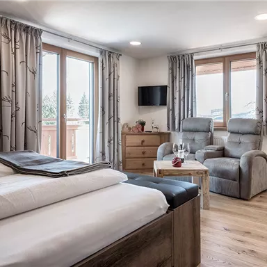 A cozy room with a double bed, a comfortable armchair, and large windows. There is a small table with glasses and a television on the wall.