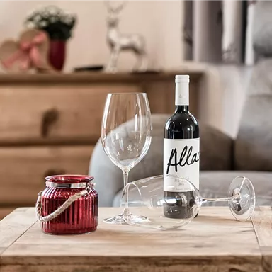 A table with a bottle of red wine, a large wine glass, and a small red tealight. In the background, a cozy armchair and other decorations can be seen.